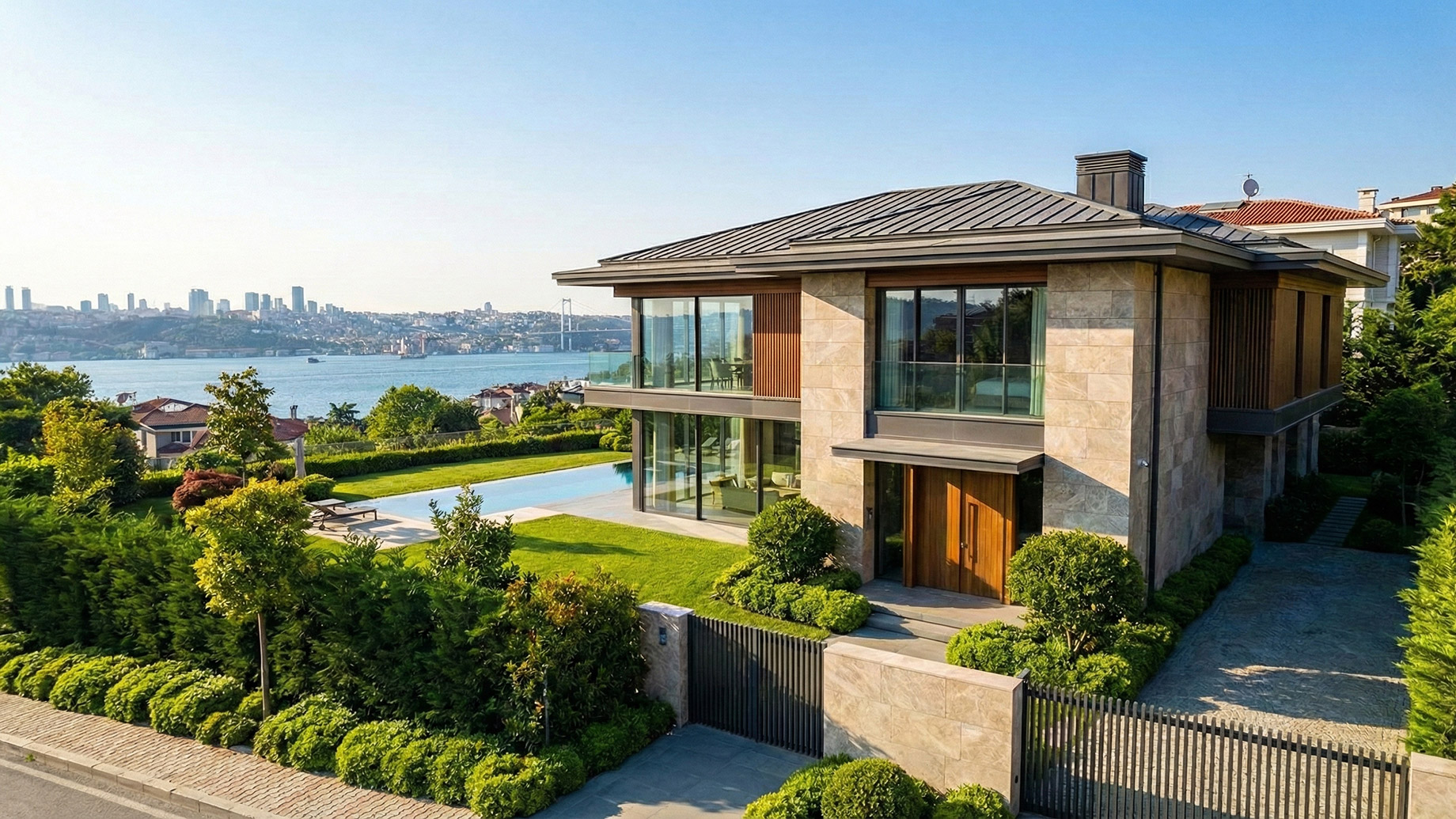 A modern, two-story luxury villa with a stone and wood facade, featuring a private swimming pool and landscaped garden. The property overlooks the Bosphorus strait with a bridge and the Istanbul city skyline in the background under a sunny sky.