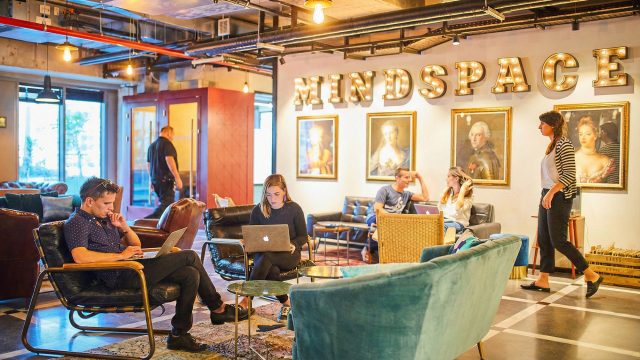 Interior view of a Mindspace flexible work environment featuring a open lounge area with the word "MINDSPACE" in large illuminated marquee letters. Professionals are working on laptops in varied seating zones that blend industrial elements with residential comfort—including velvet sofas, leather armchairs, and vintage portraits—illustrating the article's discussion on flexible zoning, layered lighting, and the shift from sterile offices to inviting, productivity-focused spaces.