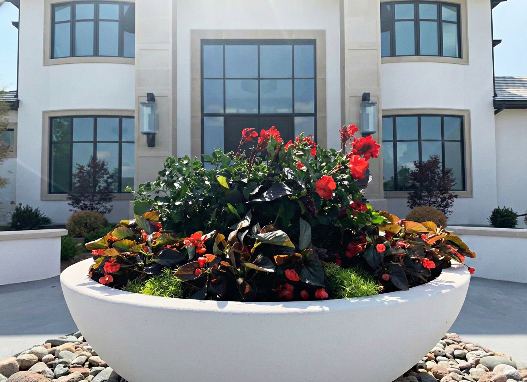 A large, modern white stone planter bowl filled with vibrant red flowers and lush greenery sits at the entrance of a contemporary commercial building. The background features a clean, white facade with symmetrical large black-framed windows, emphasizing professional landscaping and a well-maintained exterior.