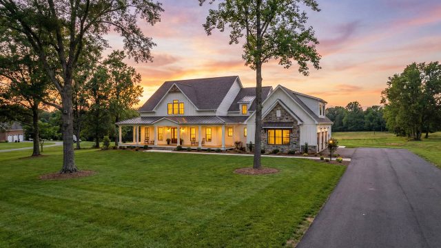 A newly constructed, two-story modern farmhouse with white siding and stone accents sits on a large, manicured lawn at twilight. The home features a wide front porch with warm interior lighting glowing through the windows, set against a colorful sunset sky, illustrating the spacious living and peaceful atmosphere available near Windsor, Virginia.