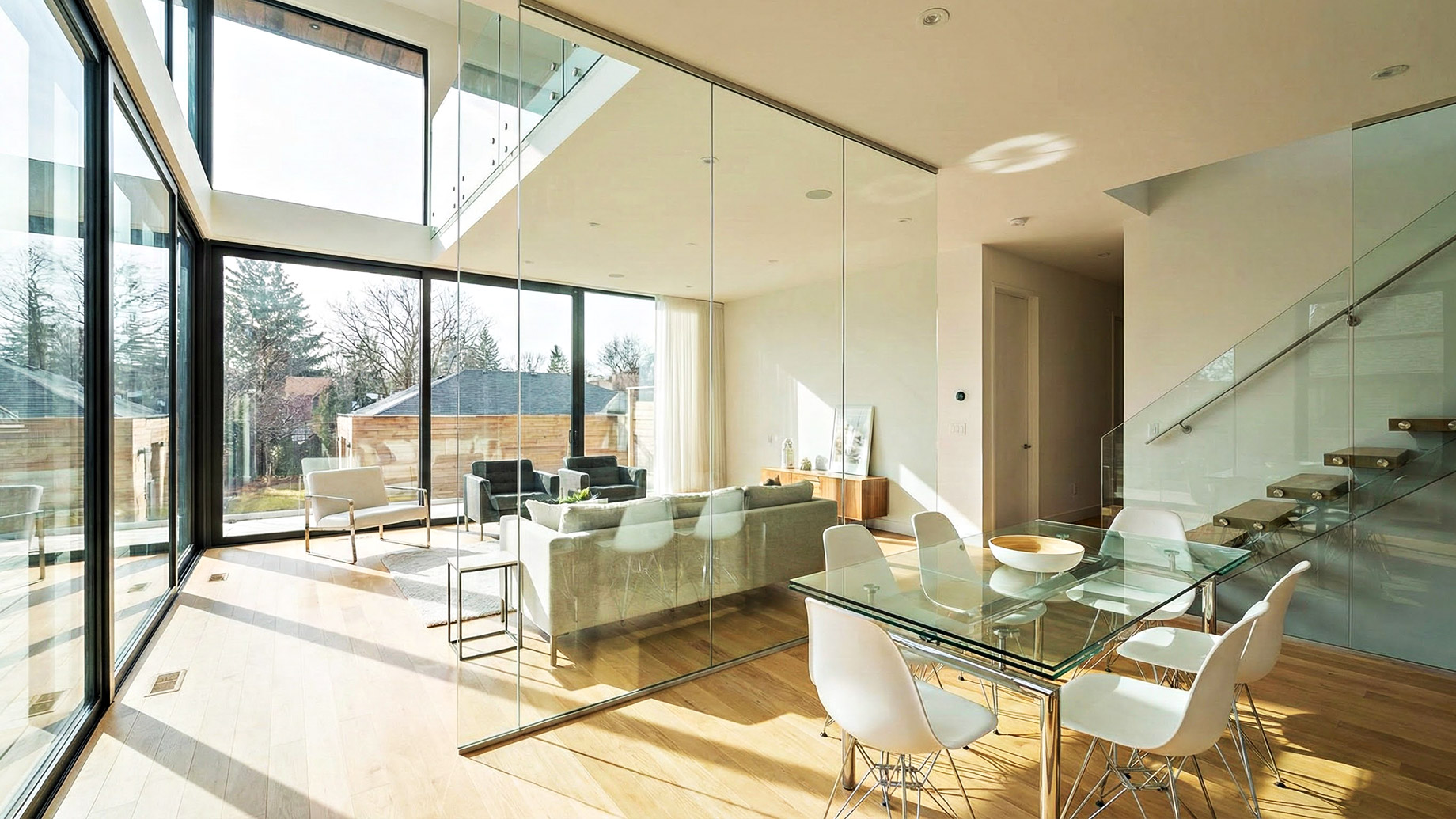 A brightly lit, modern living and dining space features large, floor-to-ceiling windows, a tall glass partition wall separating the two areas, and a staircase with a glass railing. Sunlight streams through the glass elements, creating an open and airy atmosphere with contemporary furniture and wooden flooring.