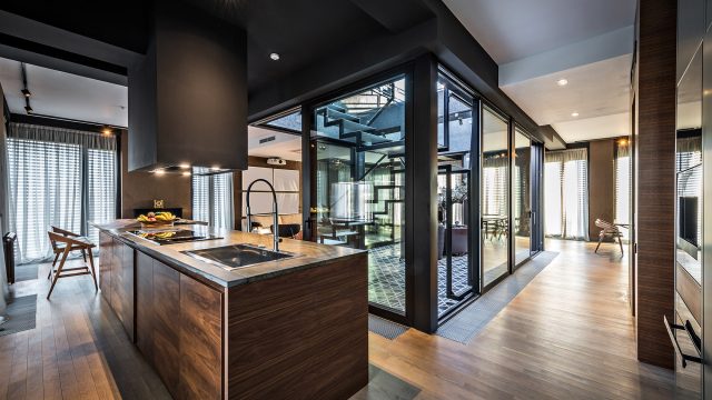 Contemporary Open-Concept Kitchen with Glass Staircase Atrium Alt Text: A sophisticated, modern kitchen and living space featuring a dark ceiling, rich walnut wood cabinetry, and a large central island with a built-in sink. The open-concept design centers around a striking glass-enclosed atrium housing a black metal staircase, with floor-to-ceiling windows and sheer curtains illuminating the warm wood flooring.