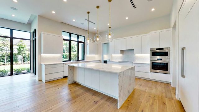 A bright, contemporary white kitchen showcasing a layered lighting scheme with three glass pendant lights over a marble island, recessed ceiling lights, and under-cabinet strip lighting.