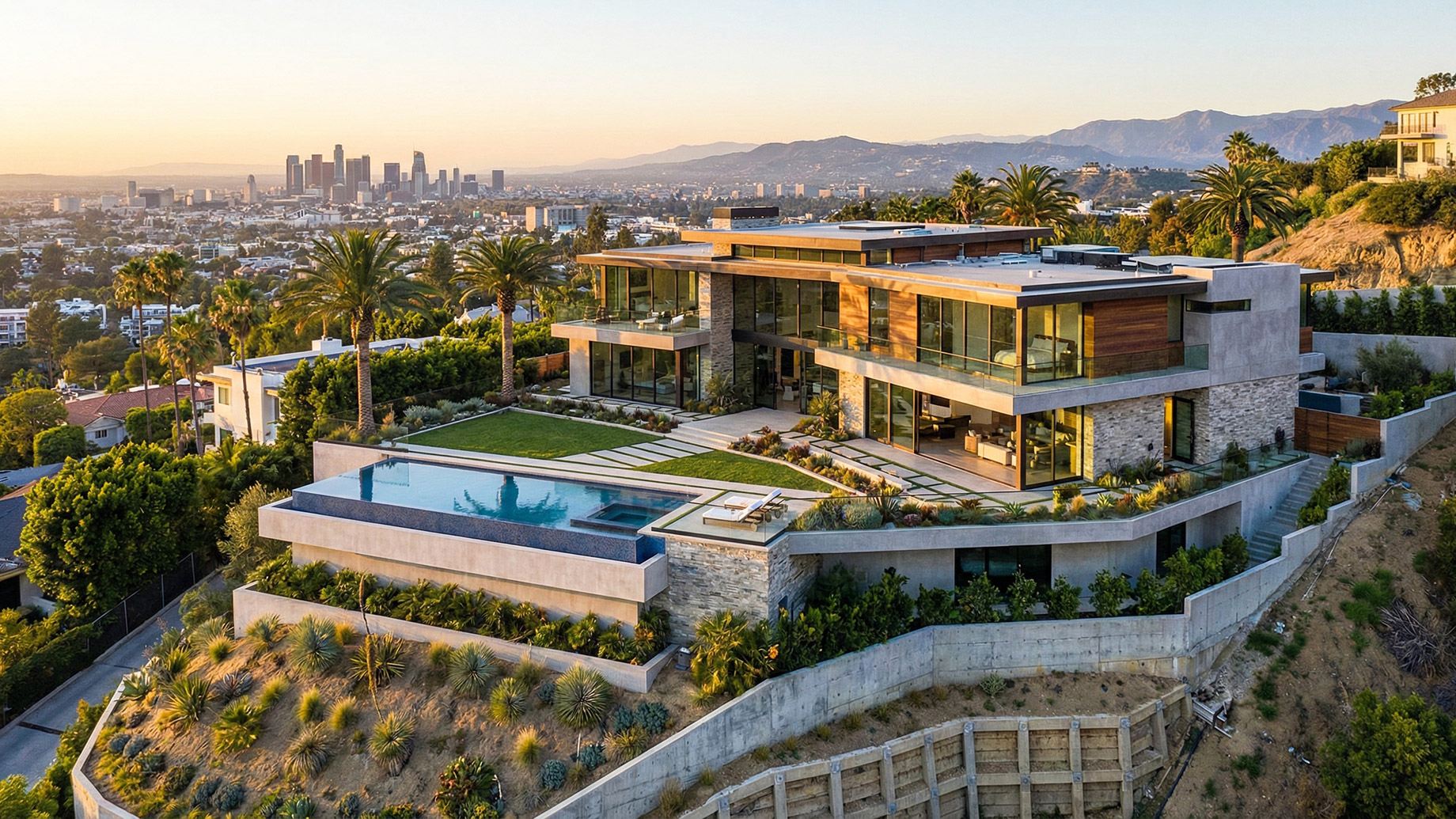 A sprawling modern luxury home perched on a steep Los Angeles hillside featuring reinforced retaining walls and a swimming pool, overlooking the downtown skyline at sunset.