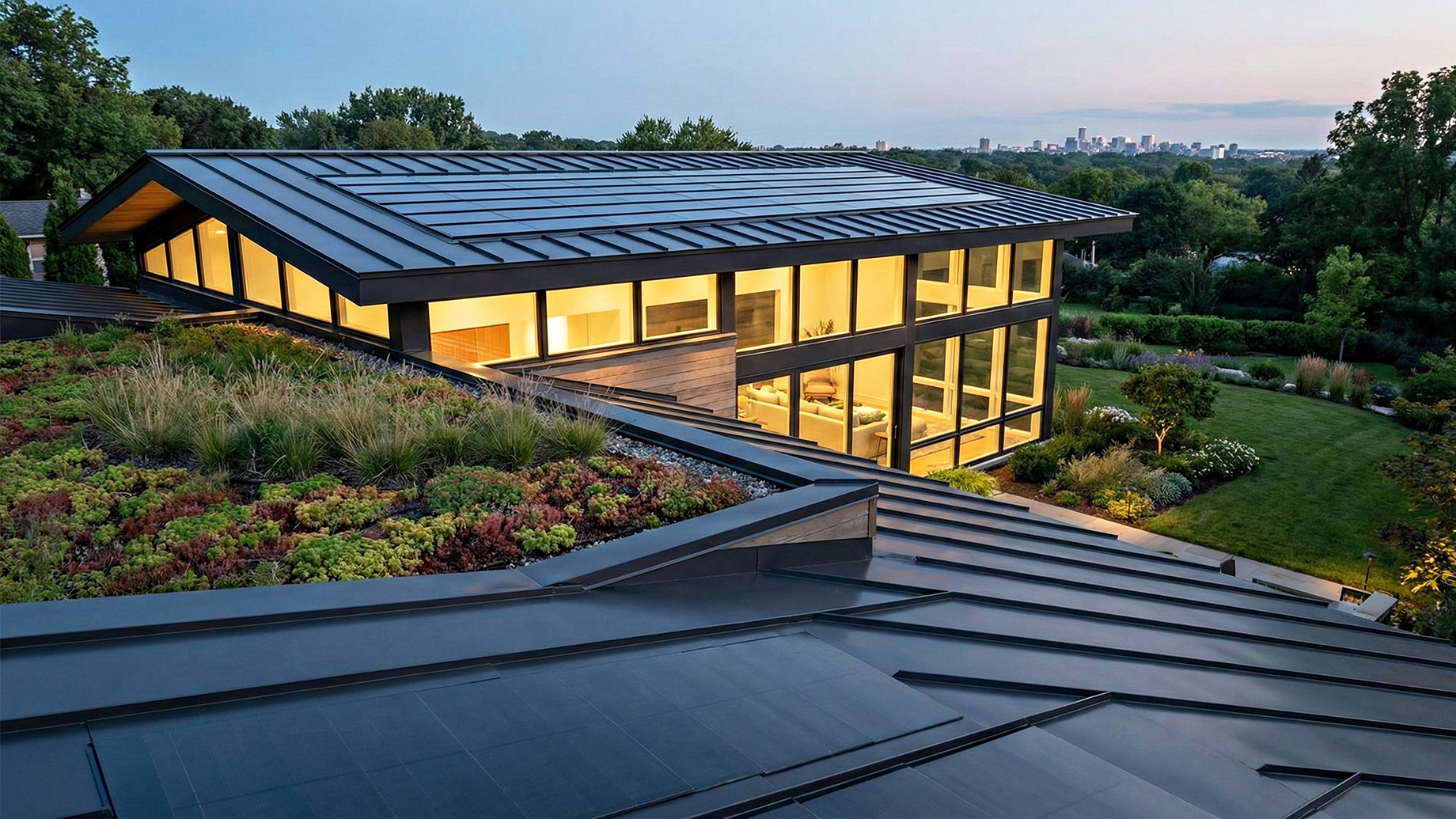 A high-angle photograph of a contemporary luxury home at twilight, showcasing advanced roofing materials. One section of the multi-level roof is a green roof covered in various low-growing plants and grasses. Another prominent section is a dark, standing-seam metal roof with solar panels integrated directly into its surface. The interior of the house is brightly lit through large glass walls, overlooking a garden and a city skyline in the distance.