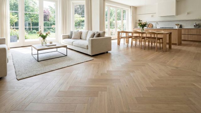 A spacious, sunlit open-concept living area featuring light neutral wide-plank hardwood flooring with a seamless transition into a custom herringbone pattern. The room showcases a matte finish floor, modern beige furniture, floor-to-ceiling windows, and a cohesive flow between the living and kitchen spaces.