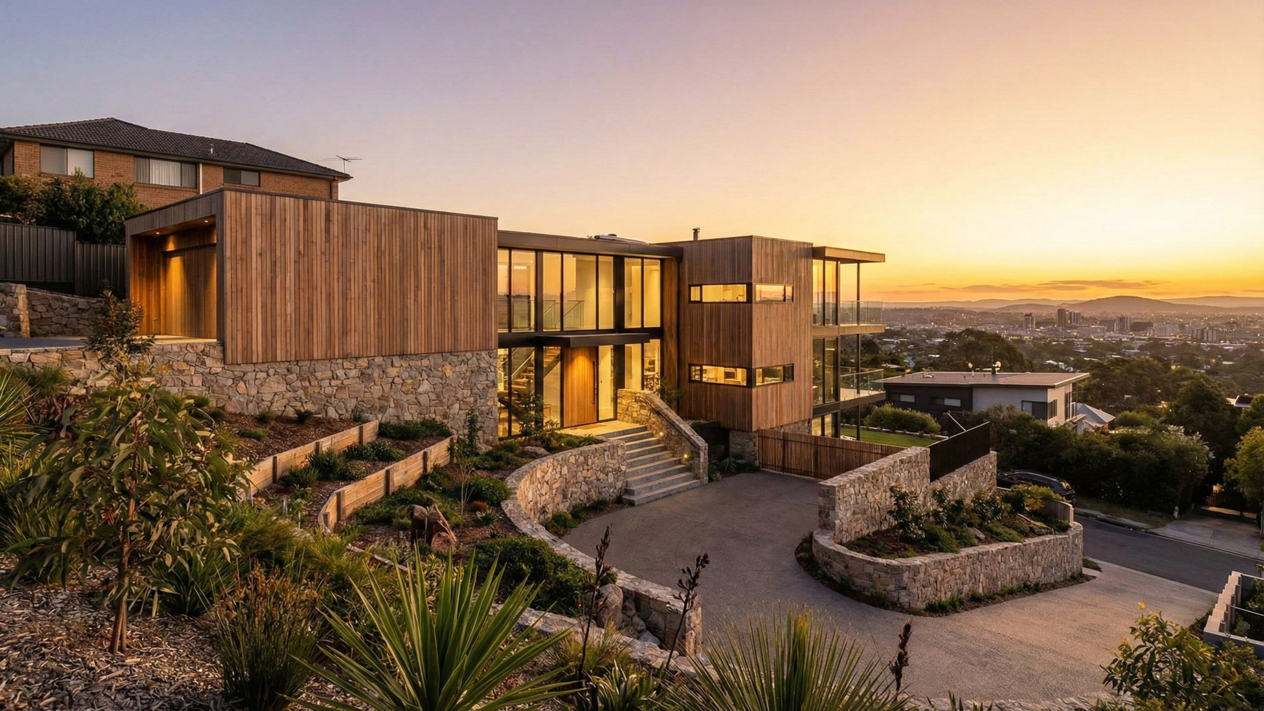 A contemporary multi-level house built on a steep hillside, featuring natural timber cladding, floor-to-ceiling glass windows, and tiered stone retaining walls. The architectural design integrates with the sloping terrain, offering a panoramic view of a city valley during a golden sunset.