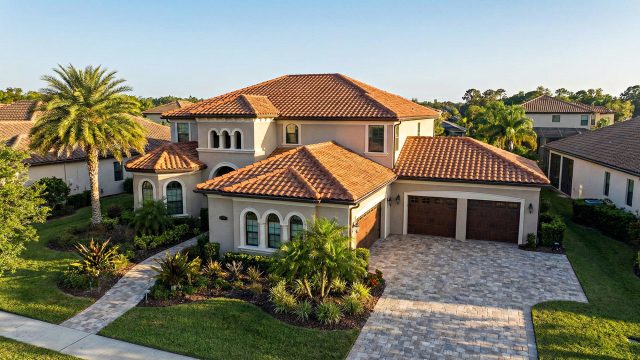 Aerial view of a large, two-story Mediterranean-style home featuring a flawless terracotta tile roof, stucco walls, and a three-car garage. A paved driveway leads up to the house, which is surrounded by manicured landscaping including a palm tree, all under a bright, sunny sky.