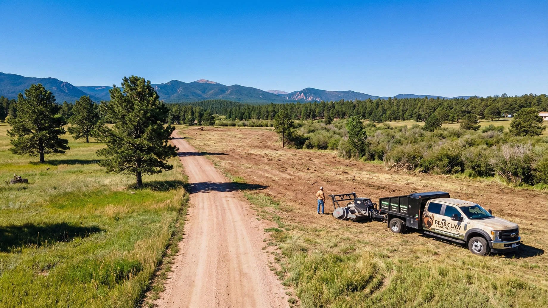 A branded Bear Claw Land Services truck with a forestry mulcher attachment is parked on a dirt road next to a person. A large section of land has been recently cleared of brush, with a forest and mountain range in the background under a clear blue sky.
