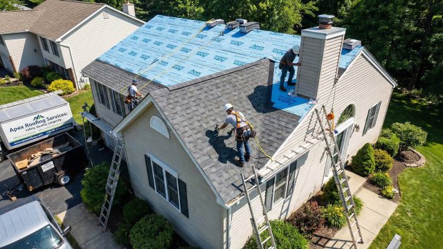 An aerial view of a professional roofing team from "Apex Roofing Services" working on a residential home. Three roofers wearing safety harnesses are on the roof, one installing new asphalt shingles over blue underlayment, while others work near a chimney. A company truck, a dumpster filled with old materials, and ladders are visible in the driveway.