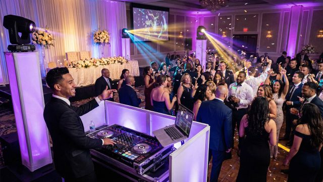 A professional wedding DJ in a formal suit mixing music and interacting with a lively crowd on a packed dance floor, surrounded by dynamic purple lighting and falling confetti at an elegant reception.