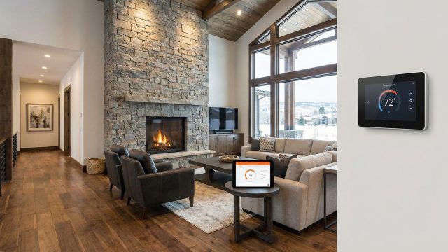 A modern smart thermostat control panel mounted on a wall displays 72° in a luxurious living room. A tablet on a side table also shows the heating controls. The room features a large stone fireplace, leather chairs, a sofa, and large windows overlooking a snowy mountain landscape.