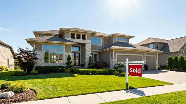 A wide-angle landscape photograph of a luxurious, modern suburban home in Kansas City on a sunny day, featuring a prominent red "Sold" sign on the well-kept front lawn.