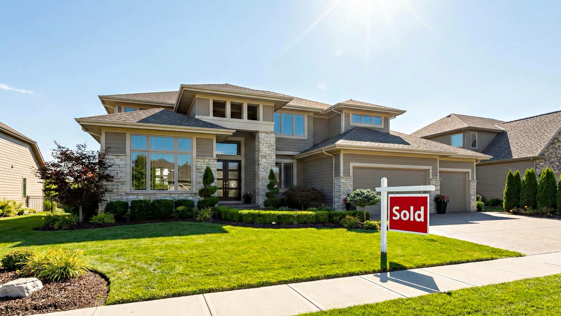 A wide-angle landscape photograph of a luxurious, modern suburban home in Kansas City on a sunny day, featuring a prominent red "Sold" sign on the well-kept front lawn.