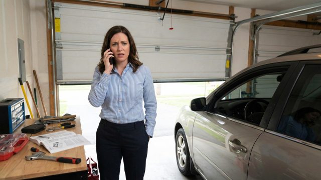 A woman with a worried expression is on the phone in her garage, standing next to a workbench with tools and a manual. Behind her, the garage door is partially open, indicating a malfunction that requires attention, possibly emergency garage door repair.