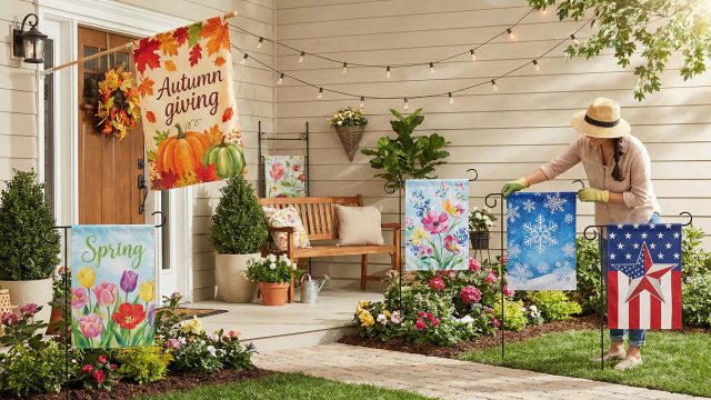 A woman in a straw hat arranges a vibrant collection of decorative garden flags in a front yard flower bed near a porch. The display features flags representing various seasons and themes, including a large "Autumn Giving" flag with pumpkins, a "Spring" tulip flag, a winter snowflake design, and a patriotic red, white, and blue star flag, all set against a cozy home exterior with hanging string lights.