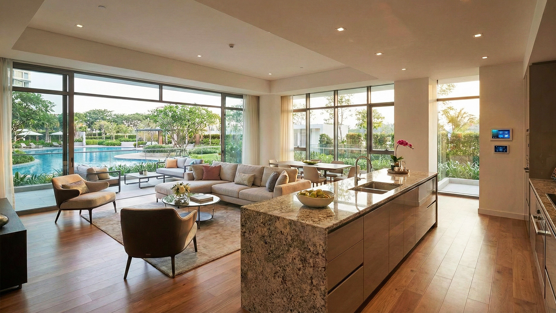 A wide-angle view of a spacious, modern luxury apartment interior. An open-plan living area with a large sectional sofa and armchairs sits next to a gourmet kitchen with a long granite island and sleek cabinetry. Floor-to-ceiling windows open onto a balcony with a view of a large resort-style swimming pool and landscaped tropical gardens under a clear sky. Hardwood floors run throughout the sunlit space.