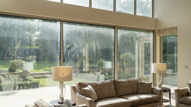 A spacious living room in a luxury home featuring floor-to-ceiling windows that are noticeably dirty with heavy streaks, haze, and dust, partially obscuring the view of the garden outside. Sunlight reveals the accumulation of grime on the glass, contrasting with the clean interior furnishings.
