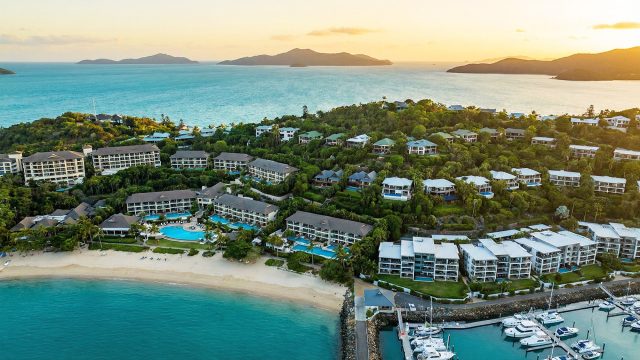 An aerial photograph taken at sunset overlooking Hamilton Island, showcasing a variety of accommodation types including large resort complexes with pools along the beachfront, private holiday homes nestled into the forested hillside, and modern apartment buildings overlooking a marina filled with boats, with the Coral Sea and distant islands in the background.