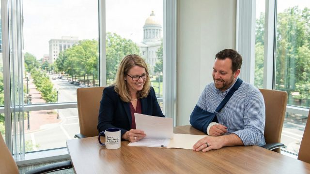 A smiling female attorney reviews a document with a male client who has his arm in a sling. They are seated at a wooden conference table with a coffee mug. Through the large office window behind them, the North Carolina State Capitol building is visible in downtown Raleigh.