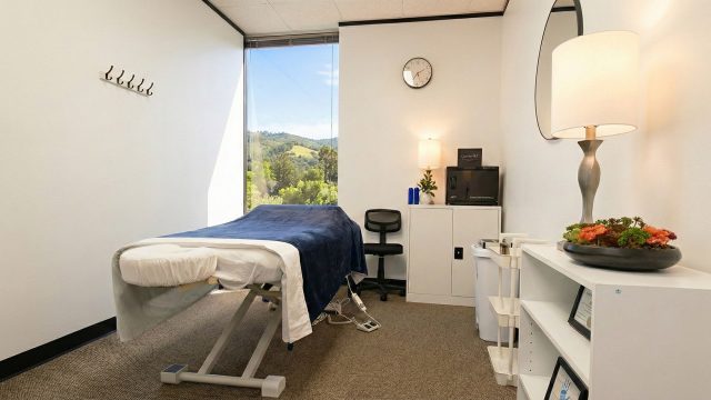 A bright, clean treatment room featuring a massage table dressed with white sheets and a blue blanket. On the left, a large, wide window provides a sweeping view of sunny, green rolling hills and trees. The room is furnished with a white storage cabinet holding a towel warmer, a black rolling stool, and a white shelving unit decorated with a lamp and a bowl of succulents.