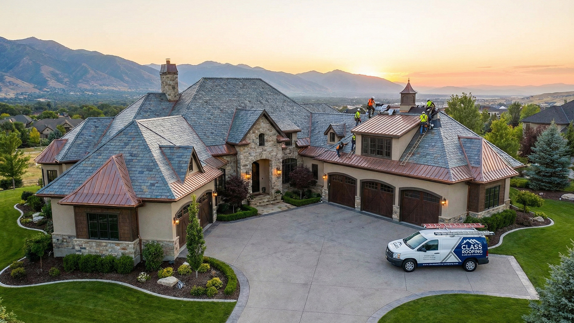 An aerial view of a large, luxury stone and stucco home with a multi-level slate and copper roof at sunset. A crew of roofers is working on a section of the roof, and a white van with the "Class Roofing" logo is parked on the large concrete driveway. Mountains are visible in the background under a warm, orange sky.
