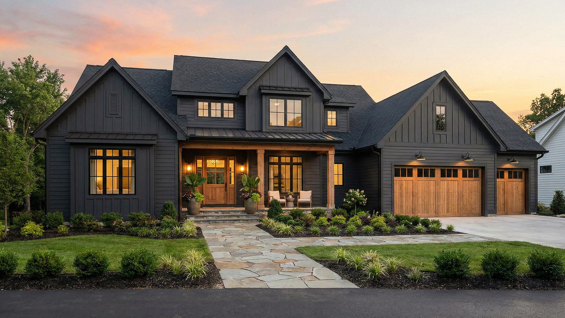 A beautiful, modern luxury home at sunset featuring dark charcoal siding perfectly contrasted with warm wood garage doors and porch columns. A natural stone pathway leads through neat, manicured landscaping to a welcoming front porch with soft exterior lighting. The consistent use of colors, black-framed windows, and natural materials creates a unified, pulled-together look.