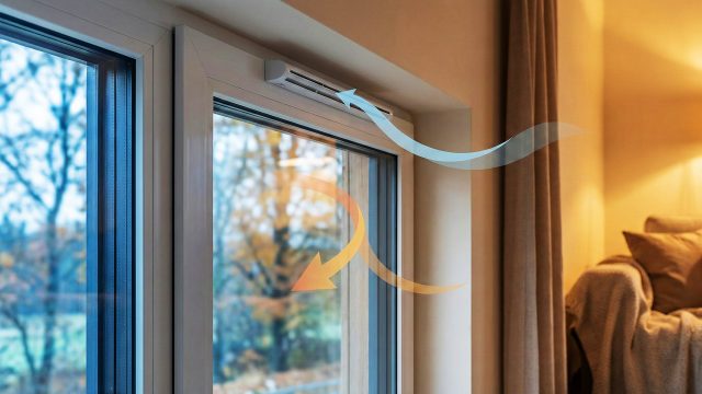 A close-up of a modern white window frame with a trickle vent installed at the top. Stylized blue arrows show cool fresh air entering through the vent, while a curved orange arrow shows warm indoor air circulating within a cozy, well-lit room with a couch and lamp. The outside view is a blurred autumn landscape.