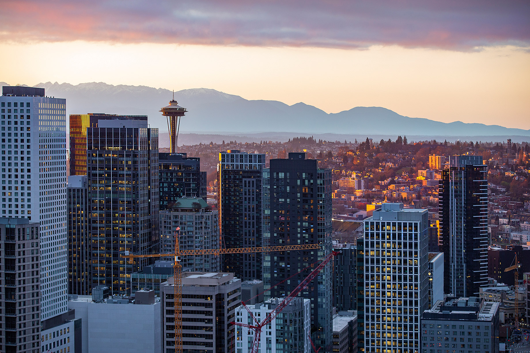 A vibrant cityscape of Seattle at dusk showing modern skyscrapers with sunlight reflecting off their windows, interspersed with construction cranes, highlighting the city's active real estate development. The Space Needle and distant mountains are visible in the background.