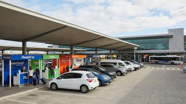 Travelers checking in at outdoor car rental kiosks at Melbourne Airport, located next to a row of parked rental vehicles ranging from compact cars to SUVs under a covered shelter, with the airport terminal and a shuttle bus visible in the background.