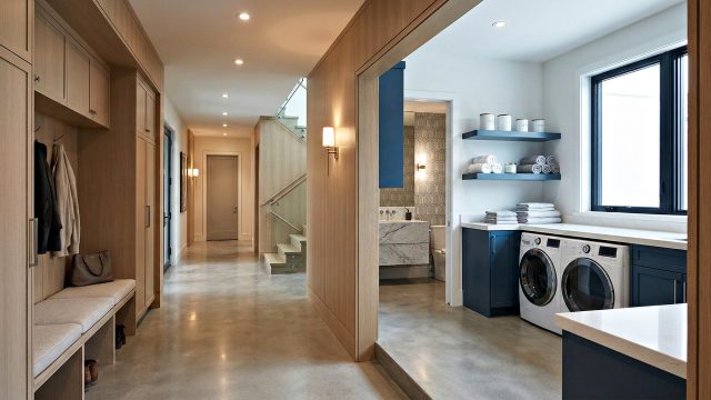 A wide view of a modern home's integrated entryway, hallway, and utility area. On the left is a custom wooden mudroom unit with a bench, coat hooks, and storage. A polished concrete hallway leads the eye past a glass-railed staircase to a distant door. To the right, a dedicated laundry room features striking blue cabinetry, white countertops, front-loading washer and dryer, open shelving with neatly folded towels, and a large window. A glimpse of a bathroom is visible behind the laundry area. Warm, integrated lighting is throughout.