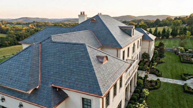 An aerial view of a luxury home featuring a complex, professionally installed slate roof with copper flashing and gutters, surrounded by a large, manicured green estate and rolling hills at sunset.