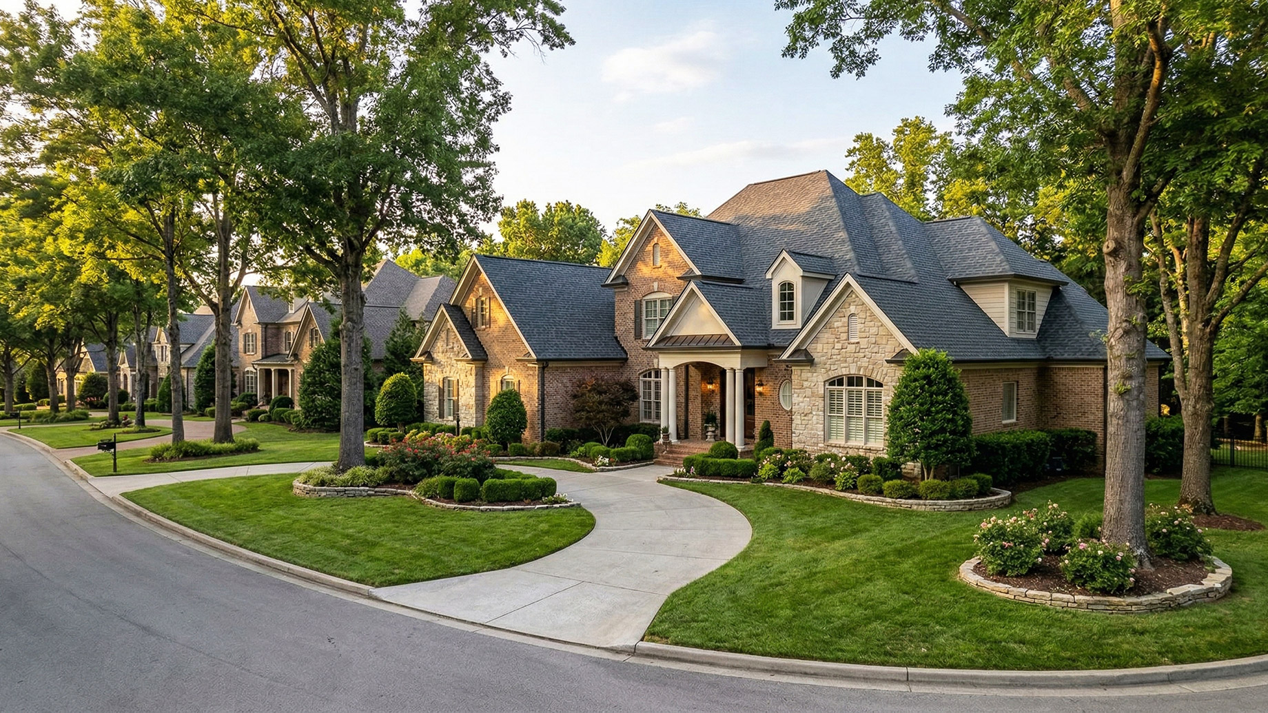 An elevated view captures a pristine, expansive traditional luxury home with a multi-gable brick and stone facade and a dark grey roof. A long, curved concrete driveway leads from a quiet, tree-lined residential street to the columned front portico. The impeccably manicured grounds feature lush green lawns, sculpted boxwood hedges, rose bushes in defined stone-edged mulched beds, and mature trees flanking the property and the street. Other upscale homes are visible along the curved road under a golden hour sky with light clouds.