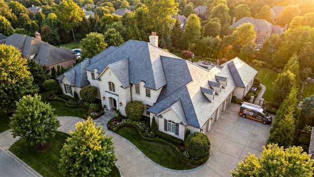 A large luxury estate with an expansive, complex gray slate roof surrounded by mature trees and manicured landscaping. A Homey Roofing branded service van is parked in the large driveway under warm evening light.