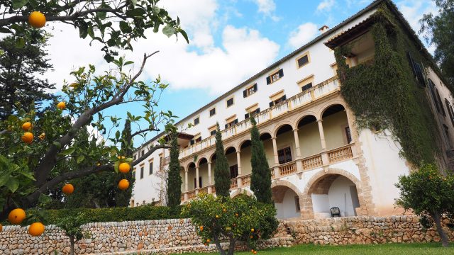 A large European luxury estate for retreats featuring classic white architecture, arched stone balconies, and beautiful grounds with a traditional stone wall and fresh orange trees.