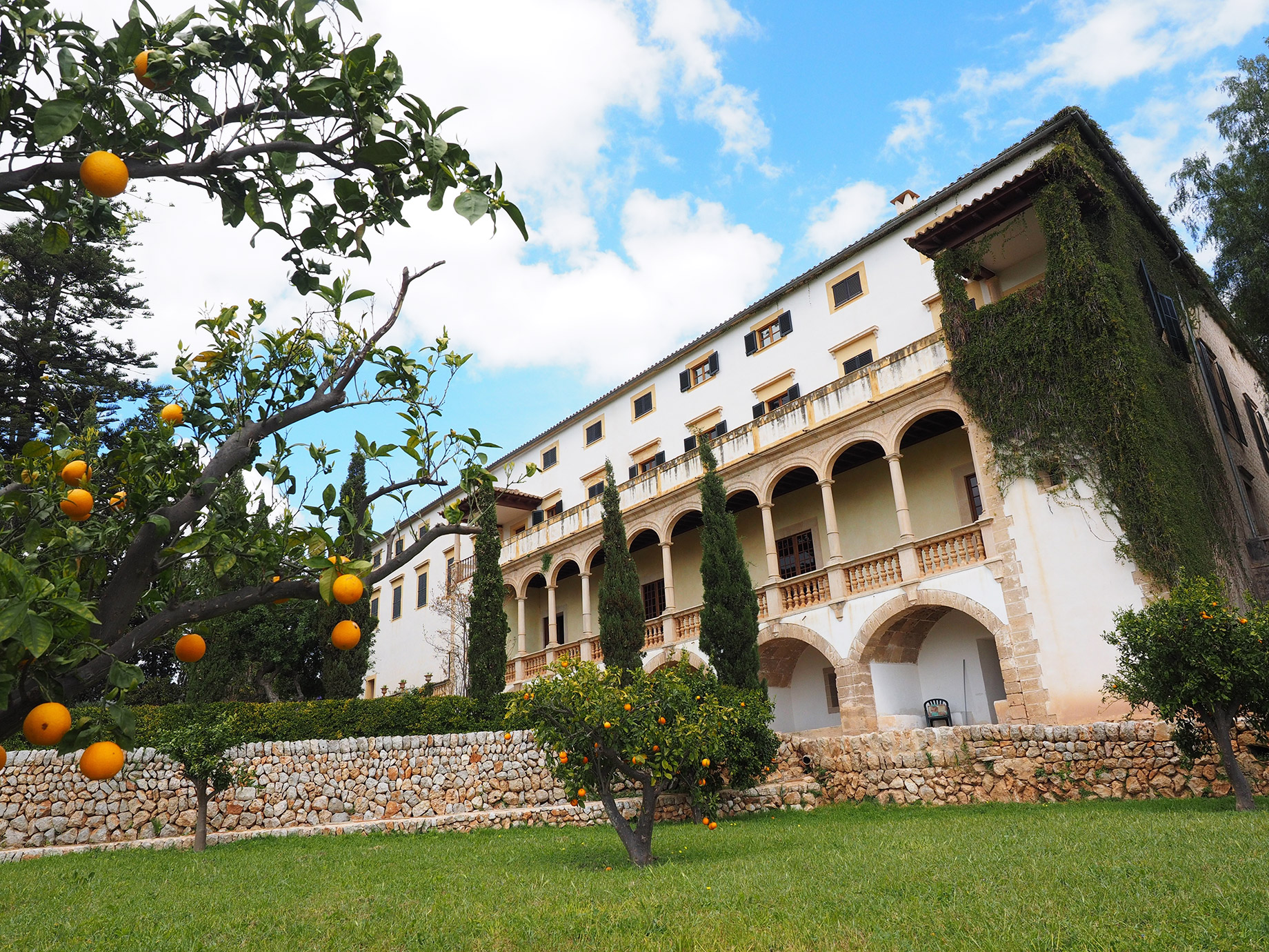 A large European luxury estate for retreats featuring classic white architecture, arched stone balconies, and beautiful grounds with a traditional stone wall and fresh orange trees.