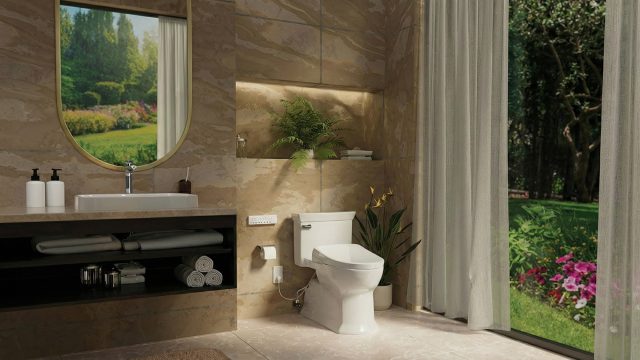 A luxurious modern bathroom featuring warm, tan stone-textured walls and flooring. On the left, a dark wood vanity holds a white rectangular sink beneath a gold-framed oval mirror that reflects an outdoor landscape. In the center sits a modern white toilet with a bidet control panel, positioned next to a tall potted plant. On the right, sheer white curtains are pulled back from a large floor-to-ceiling window, revealing a bright, lush green garden with vibrant pink flowers.