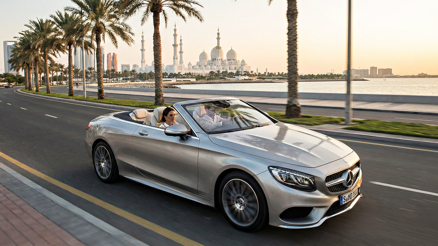 A silver Mercedes-Benz S-Class Cabriolet drives down a palm-lined coastal road in Abu Dhabi, with the Sheikh Zayed Grand Mosque in the background during sunset. A man and a woman are in the convertible, and the ocean is visible to the right.