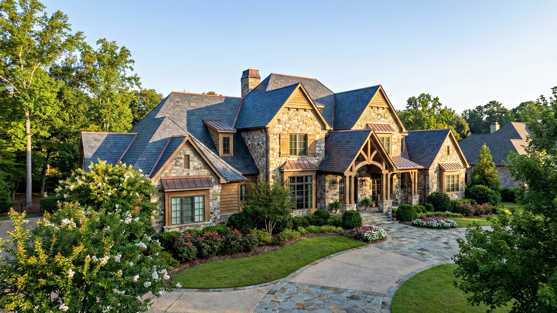 A sprawling luxury stone and wood estate featuring a pristine, multi-gabled slate roof with copper flashing accents. The home sits behind a manicured lawn, blooming bushes, and a curved stone driveway under a clear blue sky, demonstrating excellent exterior upkeep and high-end curb appeal.