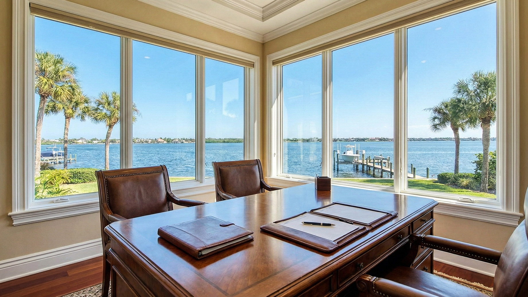 A brightly lit luxury home office featuring a large wooden executive desk and leather chairs, with expansive corner windows that offer a panoramic view of a wide river, a private dock with a boat lift, and palm trees under a blue sky.