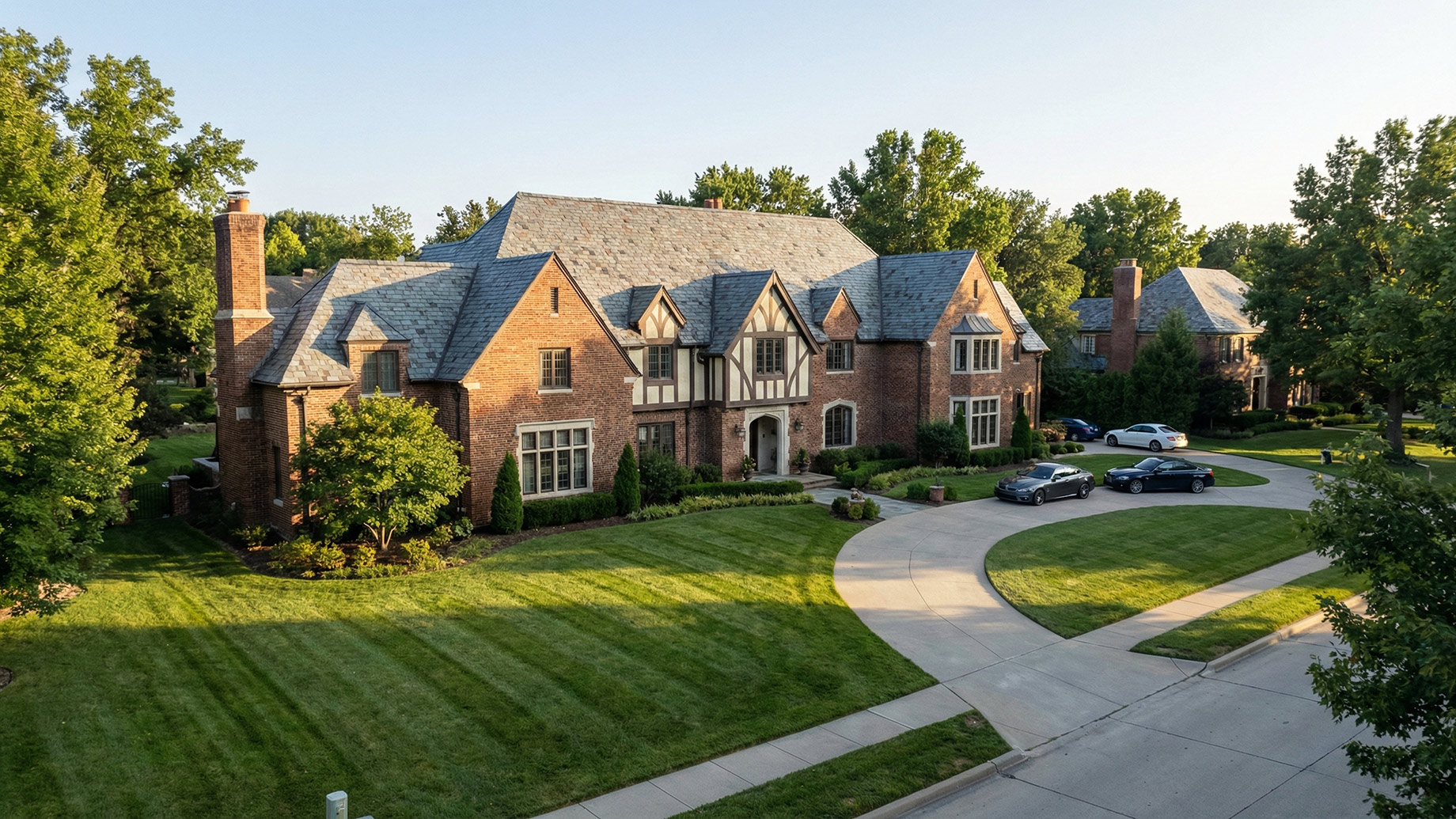A large, Tudor-style brick home with a slate roof and a well-maintained lawn in a Kansas City neighborhood, featuring a 'For Sale' sign in the front yard and three cars in the circular driveway.