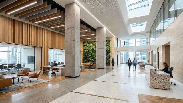 A wide-angle view of a spacious, bright multi-level modern luxury corporate office lobby. The space is characterized by exposed concrete columns, light wood slat ceiling panels with linear lighting, and large glass walls overlooking a city skyline. A prominent vertical living green wall is at the far end of the lobby. Lounge areas with modern armchairs and a conference table where people are working are present. On the right is a stone-patterned reception desk. Professionally dressed individuals walk through the open areas under large skylights.