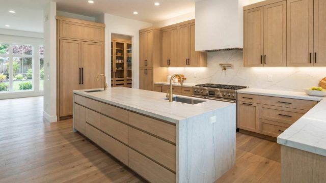 A bright, modern luxury kitchen featuring flat-panel light wood cabinetry, a massive island with a marble waterfall countertop and double brass sinks, a professional stainless steel range, and light hardwood floors. A walk-in pantry is visible in the background.