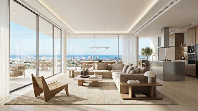 A wide-angle view of a contemporary luxury penthouse living area. A large beige sectional sofa and a wooden cane armchair sit on a textured rug facing floor-to-ceiling glass walls. In the background is a minimalist kitchen with warm wood cabinets and a stainless steel island. The expansive windows open onto a balcony with a dining table, offering a panoramic view of a coastal city skyline and the blue ocean under a clear sky.