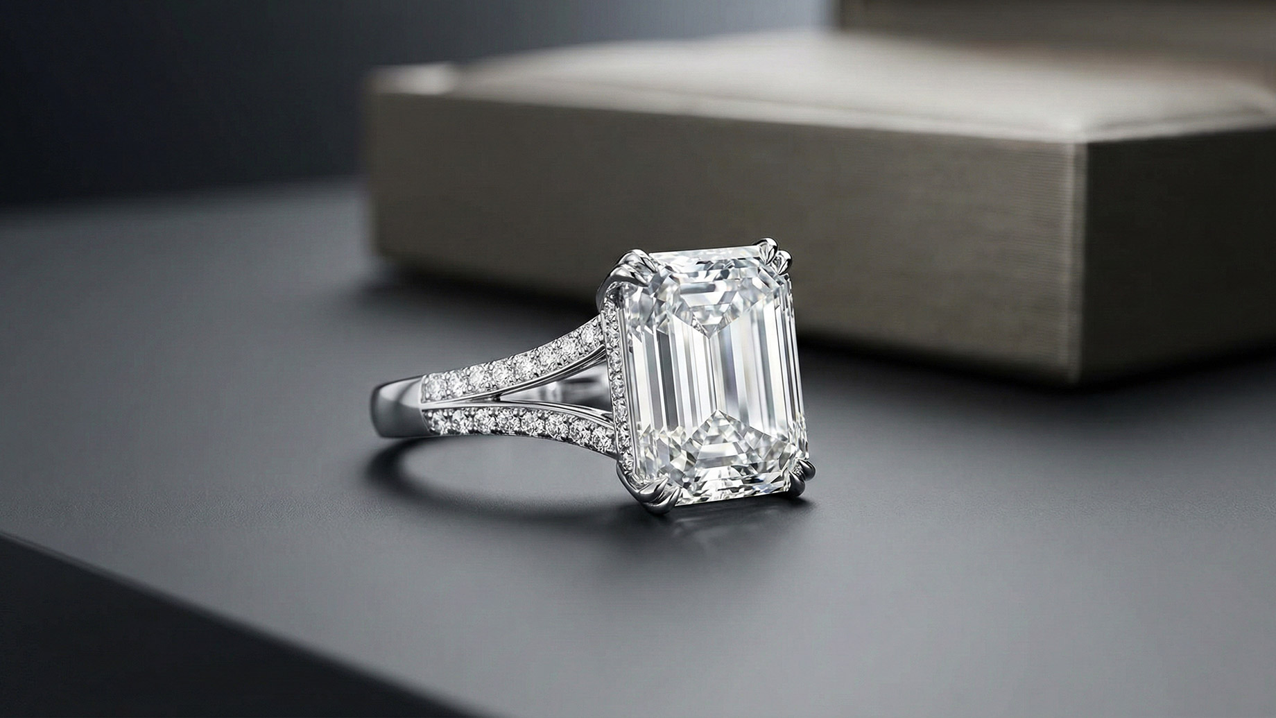 A close-up of a luxurious engagement ring featuring a large emerald-cut diamond set on a split-shank band encrusted with smaller diamonds, resting on a sleek, dark gray surface with a blurred ring box in the background.