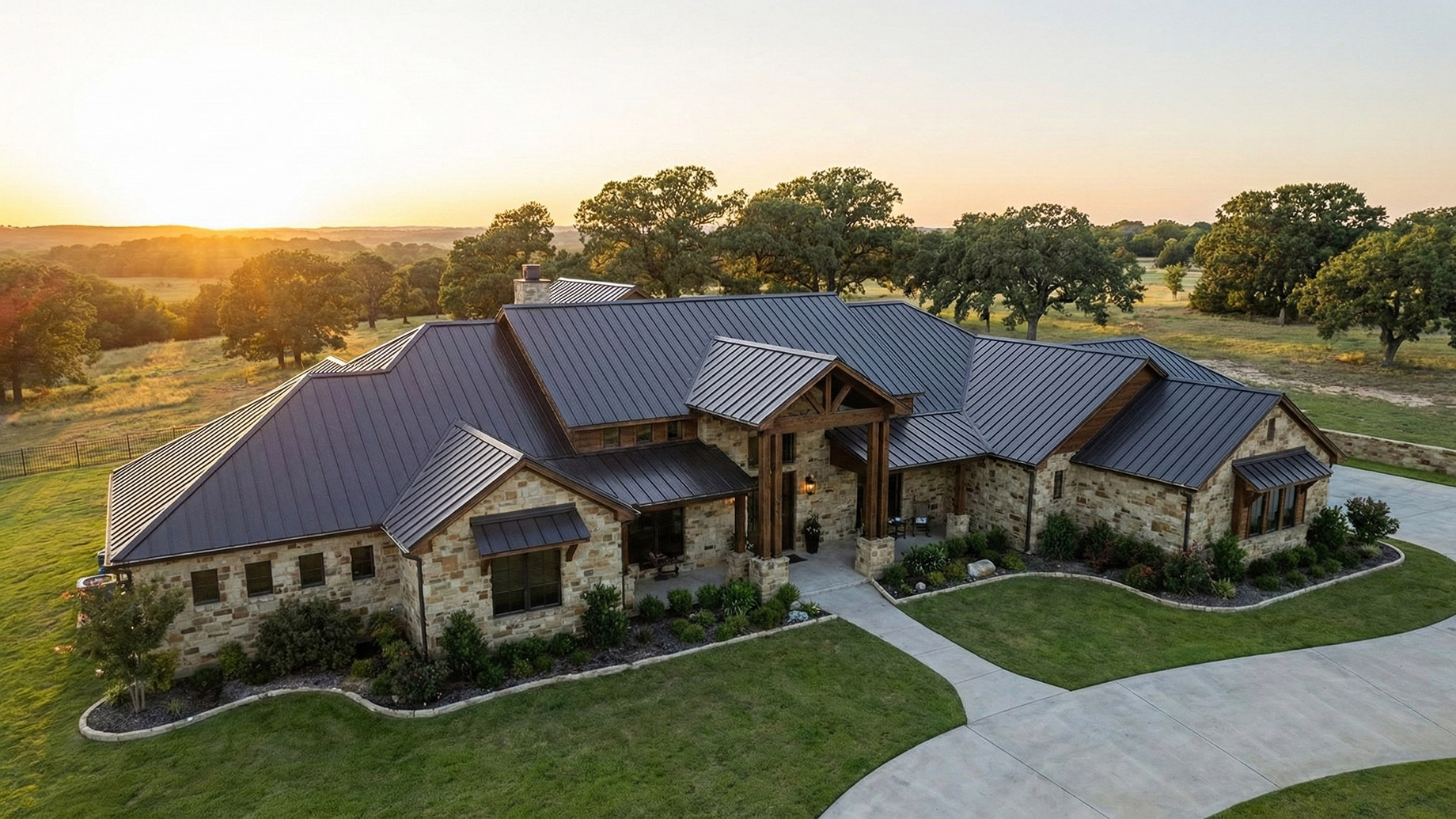 An aerial view of a sprawling luxury stone estate featuring a sleek, dark standing-seam metal roof at sunset. The home is surrounded by a manicured lawn and mature trees, highlighting the durability and aesthetic appeal of modern metal roofing in a rural setting.