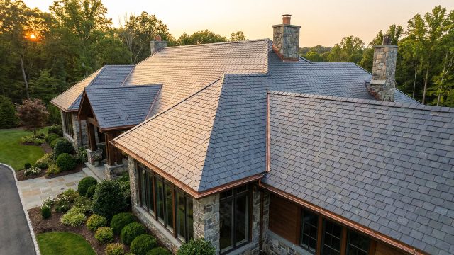 A high-angle view of a large, multi-gabled slate roof with copper gutters on a luxury stone and wood home, glowing in the warm light of sunset, illustrating the value of proactive maintenance for long-term structural integrity.