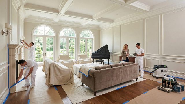 A spacious luxury living room being prepared for professional painters. Furniture is carefully covered with drop cloths and moved to the center of the room to clear the walls. A female homeowner consults with a lead painter holding a tablet, while another crew member applies blue painter's tape to a fireplace mantel, illustrating the initial stage of a high-end painting project.