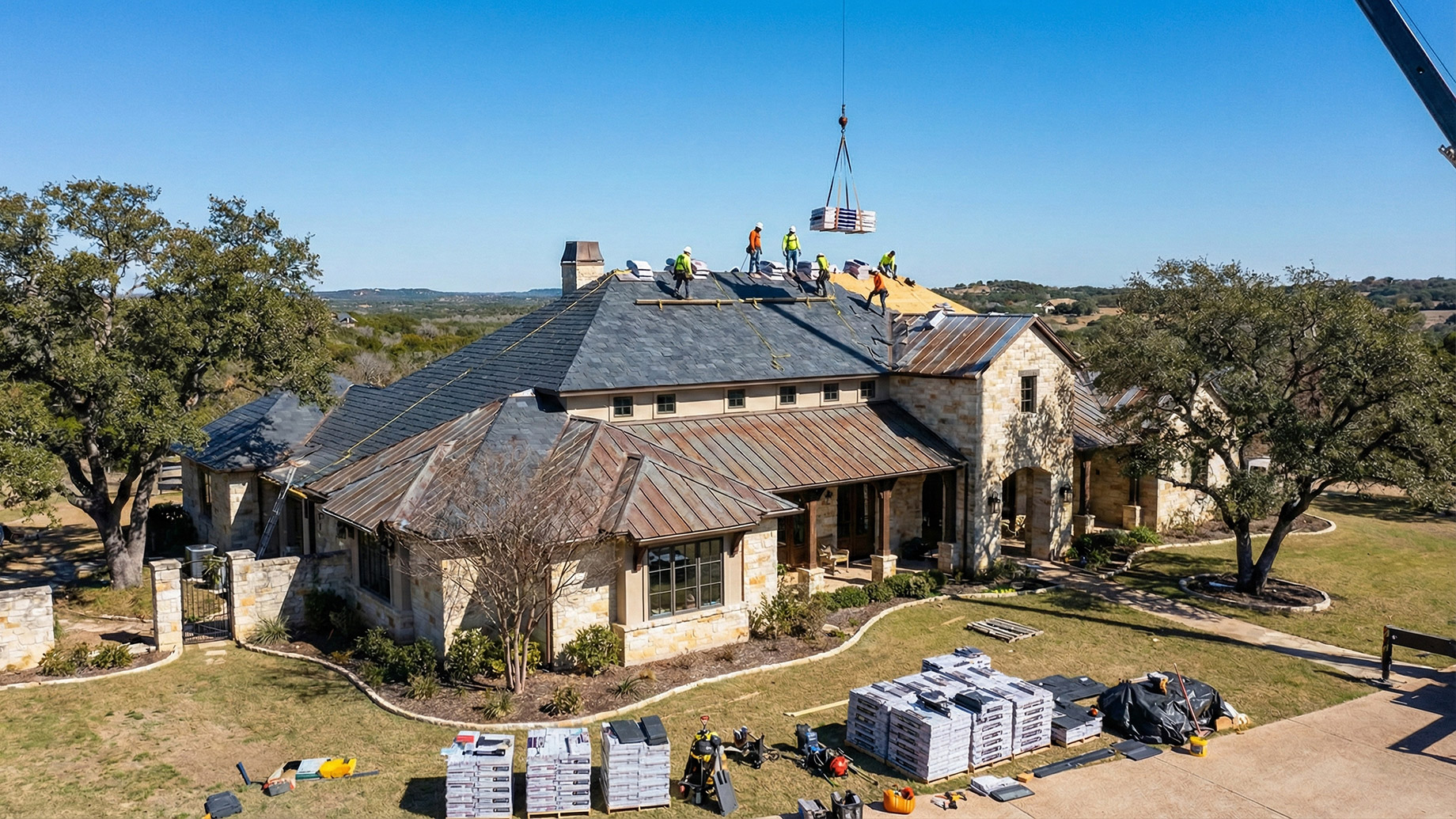 An aerial view shows a crew of workers replacing the slate roof on a large stone house. A crane is lifting roofing materials to the roof, and stacks of new materials are on the lawn below. The house is surrounded by trees under a clear blue sky.