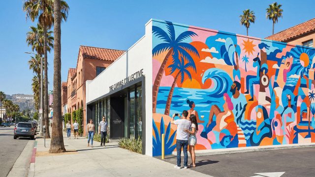 A sunny street-level view of a modern luxury business in Los Angeles featuring a large, colorful exterior mural with abstract tropical themes. A couple stops to take a photograph of the artwork while other pedestrians walk past, demonstrating how street art attracts attention and increases foot traffic.